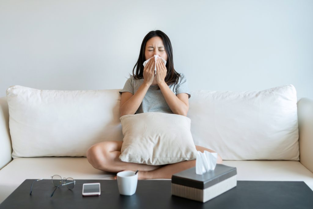 Sick young woman sitting on couch blowing her nose on a tissue conceptual of healthcare , seasonal flu, rhinitis or an allergic reaction in hay fever.