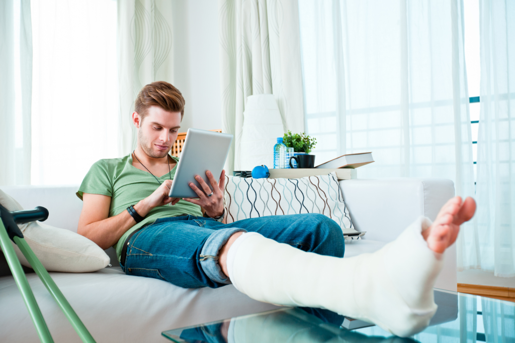 Man is sitting on couch with a cast on his right foot and is using a tablet.
