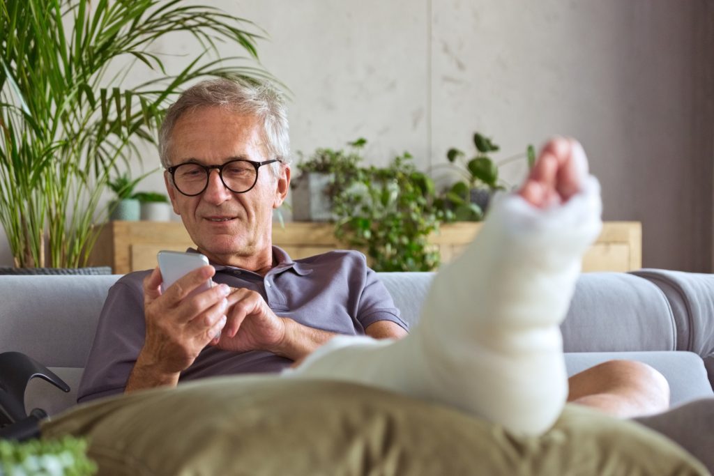 Cheerful senior man with broken leg in plaster cast sitting on sofa at home and using smart phone.